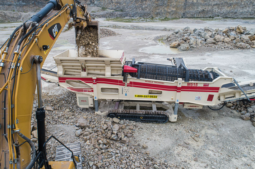 An excavator dumps a bucket of aggregates into a trommel.